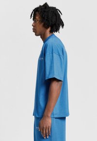 Sininen puuvillainen ylisuuri t-paita, jossa on lyhyet hihat, pyöreä kaula-aukko ja etupuolella logo, yhdistettynä samansävyisiin sinisiin shortseihin. Sujuva pinta.