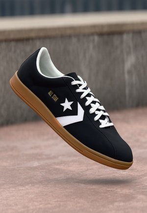Black low-top sneaker with white star and chevron logo, white laces, "All Star" text, and gum rubber sole, floating above a concrete surface.