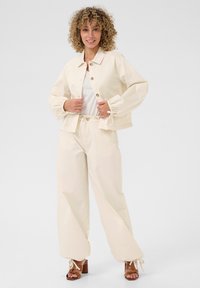 Beige cropped jasje met knopen aan de voorkant, met pofmouwen; gecombineerd met bijpassende losse broek met strik in de taille en bruin met hoge hakken sandalen.
