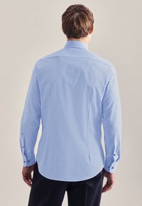 Camicia azzurro chiaro con maniche lunghe, polsini abbottonati e orlo curvo. Tessuto liscio e resistente alle pieghe con una leggera lucentezza.