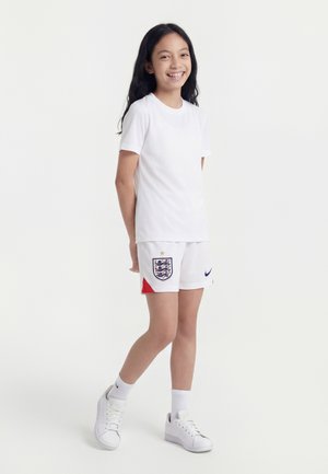 Fille souriante debout en tenue de sport blanche avec l'écusson de football d'Angleterre sur le short et des baskets blanches sur fond uni.