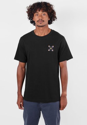 Homme aux cheveux bouclés portant un T-shirt noir avec un petit motif floral coloré sur le côté gauche de la poitrine et un pantalon bleu sur un fond uni.