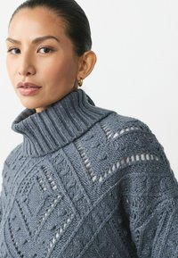 Grå stickad tröja med hög ribbad krage, med texturerade mönster och öppna stygn genom hela tyget.