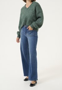 Grüner, grob gestrickter Pullover mit V-Ausschnitt; kombiniert mit hoch taillierter, weit geschnittener blauer Jeans und schwarzen Absätzen. Die strukturierte Stoffoberfläche kontrastiert mit dem glatten Denim.