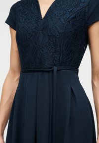 Μπλε φόρεμα ναυτικού με κορσάζ από δαντέλα, κοντά μανίκια, ντεκολτέ σε σχήμα V, ζώνη στη μέση και φαρδιά φούστα σε όρθιο άτομο.