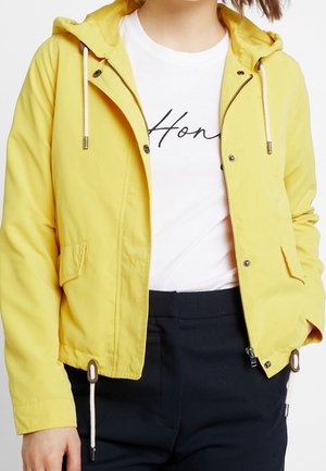 Veste à capuche jaune avec fermeture éclair et boutons-pression portée sur un t-shirt blanc avec le texte manuscrit "Hon", associée à un pantalon foncé.