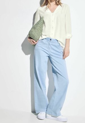 Femme portant un pantalon large bleu clair, une chemise crème à boutons, des baskets blanches, tenant une pochette verte texturée sur un fond blanc.