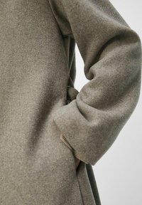 Abrigo de lana en gris verdoso apagado, con un corte relajado, bolsillos laterales y costuras visibles a lo largo de los bordes. Se muestra una tela suave y texturizada.