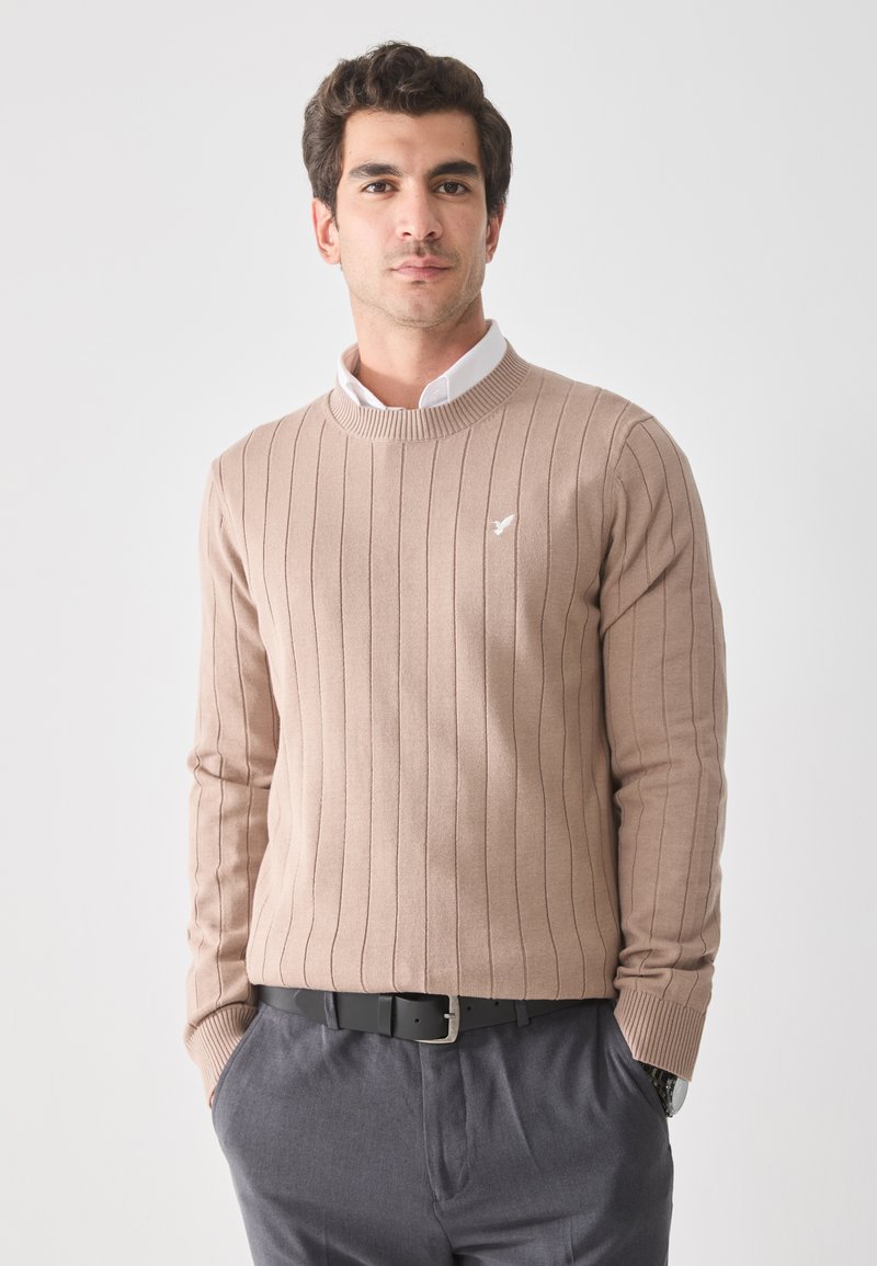 Pull en maille côtelée brun clair avec un col rond, arborant un petit logo blanc sur la poitrine, associé à un pantalon gris et une ceinture noire.