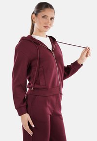 Felpa con zip color bordeaux, dotata di cappuccio con cordino, tasche laterali e polsini a coste. Realizzata in materiale morbido, abbinata a pantaloni della tuta coordinati.