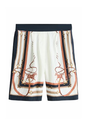 Person iført hvide shorts med mønster af hest, reb og bælte står nær en flisebelagt fontæne udendørs.