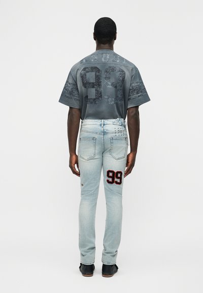 Άνδρας που φοράει ξεθωριασμένο μπλε τζιν με μπαλώματα αριθμού 99 στο μηρό και γκρι μπλουζάκι με τον αριθμό 99 στην πλάτη, στεκόμενος με την πλάτη γυρισμένη.