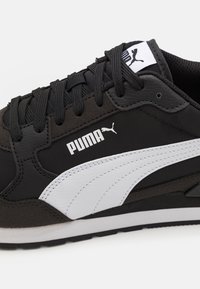 Zapatilla negra con materiales lisos y texturizados, que cuenta con una franja blanca lateral y un logo, cordones redondos y una suela de goma plana con agarre.