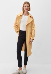 Femme portant un trench-coat jaune pâle sur une chemise blanche, un pantalon slim foncé et des chaussures blanches à enfiler, se tenant devant un fond blanc.