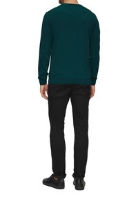 Dunkelgrüner Pullover mit langen Ärmeln und rundem Ausschnitt, kombiniert mit schwarzen Hosen und schwarzen Schuhen. Der Stoff wirkt weich und strukturiert.
