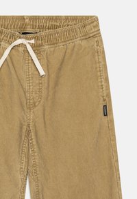 Kordfløyelsbukser i beige med snor i midjen og sidelommer. Tekstilt stoff med vertikal ribbing, inkludert en merkeetikett.
