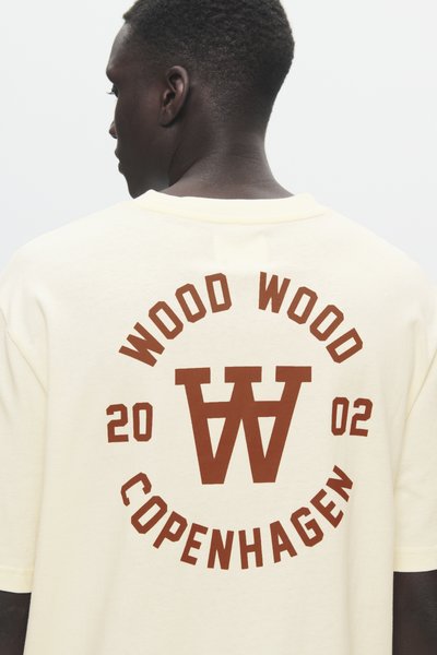 Persona con capelli corti e ricci che indossa una camicia cremosa con la scritta rossa "WOOD WOOD 2002 COPENHAGEN" sulla schiena e un logo stilizzato a doppia W.