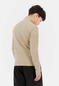 Beige fleece pullover met een hoge kraag, lange mouwen en een gladde textuur, gedragen door een persoon die wegkijkt. Zwarte broek is zichtbaar.