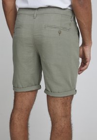 Pantalones cortos verde oliva de tejido ligero, con dobladillos enrollados y dos bolsillos traseros con cierre de botón. Textura suave, diseño casual.