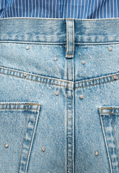 Οπίσθια όψη ανοιχτού μπλε τζιν με ασημένιες τρουκς και πορτοκαλί ραφές, φορεμένο με μπλε και λευκή ριγέ πουκάμισο.