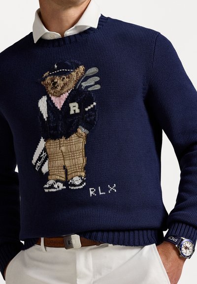 Maglione lavorato a maglia blu navy con un orso ricamato in abbigliamento da golf, pantaloni a quadri marroni e colletto rosa. Include il logo "RLX" nella parte inferiore.