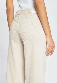 Beige corduroy pants with wide legs, featuring two back pockets and visible stitching detail. The waistband is textured with a label.
