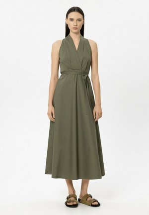 Frau in ärmellosem olivgrünem Wickelkleid mit gebundenem Gürtel und braunen Sandalen mit offenen Zehen, stehend vor einem einfarbigen weißen Hintergrund.