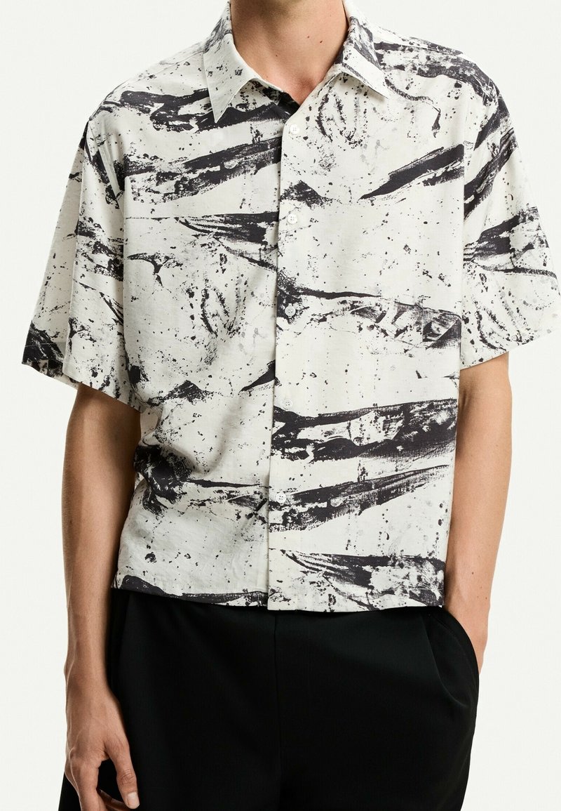 Lyhythihainen paita kermaisenvärisestä kankaasta, jossa musta abstrakti kuvio. Mallissa on nappikiinnitteinen kaulus ja rento istuvuus.