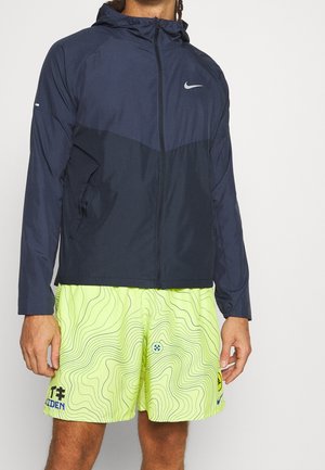 Male wearing navy blue Nike hooded zip jacket and neon yellow athletic shorts with black topographic lines and logos, standing against white background.