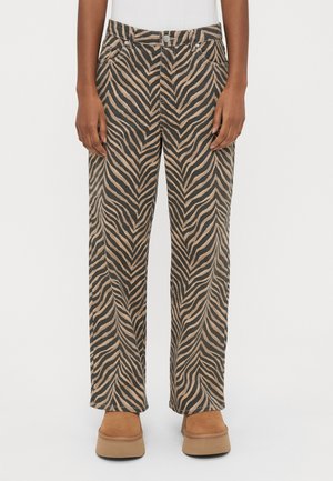 Person trägt braune und schwarze, zebra-gestreifte Hosen mit weitem Bein, beige Plateau-Schuhe und weißes Oberteil, steht vor einfachem Hintergrund.