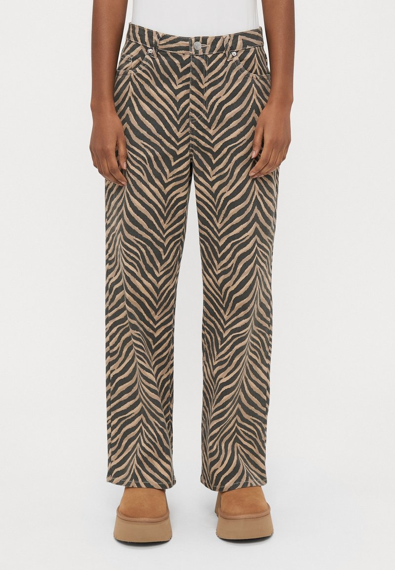 Personne portant un pantalon large à rayures zèbre marron et noir avec des chaussures à plateforme beige et un haut blanc, debout sur un fond uni.