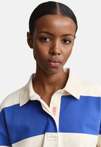 Jeune femme à la peau lisse et aux cheveux tirés en arrière, portant une chemise crème et bleue à col avec une étiquette rouge visible.