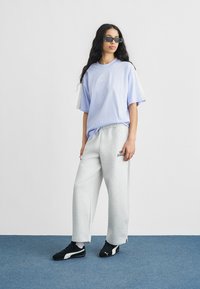 Femme aux longs cheveux foncés portant des lunettes de soleil, un T-shirt ample violet clair, un pantalon de survêtement gris à jambes larges et des baskets Puma noires, debout sur un sol bleu.