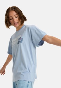 Jasnoniebieska, oversize t-shirt z bawełny, z kwiatowym wzorem i napisem "ŁAMIĄC SERCA". Krótkie rękawy i luźny krój.