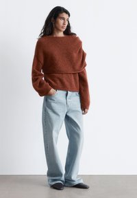 Rust-colored knit sweater with a draped design, paired with light blue wide-leg jeans and black slip-on shoes. Minimalist style.