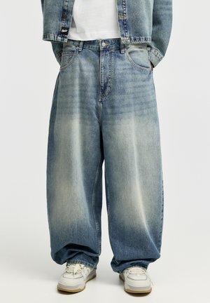 Person trägt übergroße ausgebleichte blaue Jeans, weiße Sneakers, weißes Hemd und teilweise sichtbare Jeansjacke mit den Händen in den Taschen.
