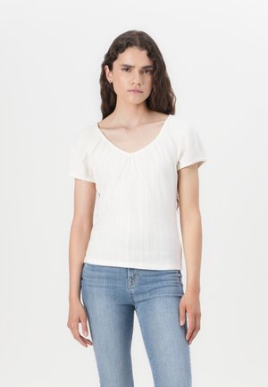 Haut blanc à manches courtes avec un col en V, présentant des rayures verticales texturées et une coupe ajustée. Associé à un jean bleu pour un contraste.