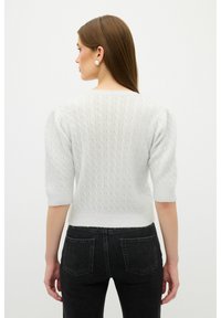 Weißer Strickpullover mit kurzen Puffärmeln und strukturierten Rautenmuster. Kombiniert mit hochgeschnittenen dunklen Jeans. Schlichtes und modernes Design.