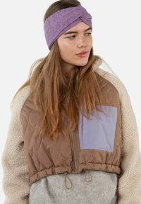 Veste courte en cuir matelassé marron avec une poche violette et des manches en polaire crème ; accompagnée d'un bandeau torsadé lavande. Textures douces évidentes.