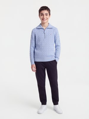 Băiat zâmbitor în picioare, purtând un pulover bleu deschis cu model împletit și fermoar până la un sfert, pantaloni negri și adidași albi, pe un fundal simplu.