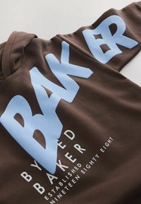 Hnědá mikina s velkým světle modrým textem "BAKER" a menším bílým textem "BY RED BAKER ESTABLISHED NINETEEN EIGHTY EIGHT" pod ním.