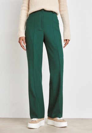 Henkilö, jolla on ylhäälle korkeavyötäröiset, leveälahkeiset tummanvihreät housut, beigejä paksupohjaisia tasapohjasneakers-kenkiä ja kermaan vivahtava neulepaita.