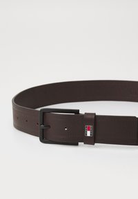 Cintura in pelle marrone con finitura testurizzata, design piatto e fibbia in metallo nero con una patch logo con accenti rossi, bianchi e blu.