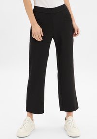 Pantalon large noir en tissu lisse, avec deux poches avant, conçu avec une taille haute pour le confort. Assorti à des baskets blanches.