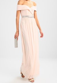 Robe longue rose clair à épaules dénudées, avec un corsage ajusté et une jupe fluide. Dotée d'une ceinture ornée de bijoux et d'une paire de talons ouverts argentés.