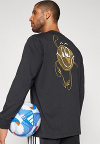 Camisa negra de manga larga con un diseño de dibujos animados en dorado en la espalda. El modelo sostiene un balón de fútbol de color azul y blanco con estampado.