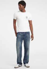 Witte t-shirt met grijze accenten, voorzien van een logo, gecombineerd met versleten blauwe jeans en witte sneakers met zwarte strepen. Man staat casual.