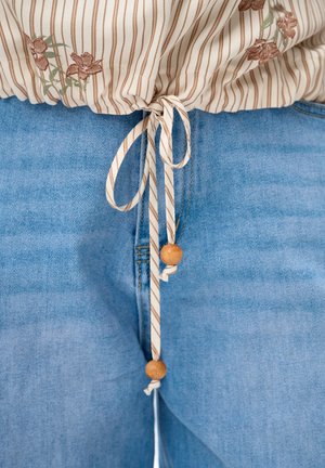 Beżowy top w paski z kwiatowym haftem, zawiązany w talii, oraz jasnoniebieskie dżinsy widoczne pod węzłem, z drewnianymi koralikami na sznurkach.