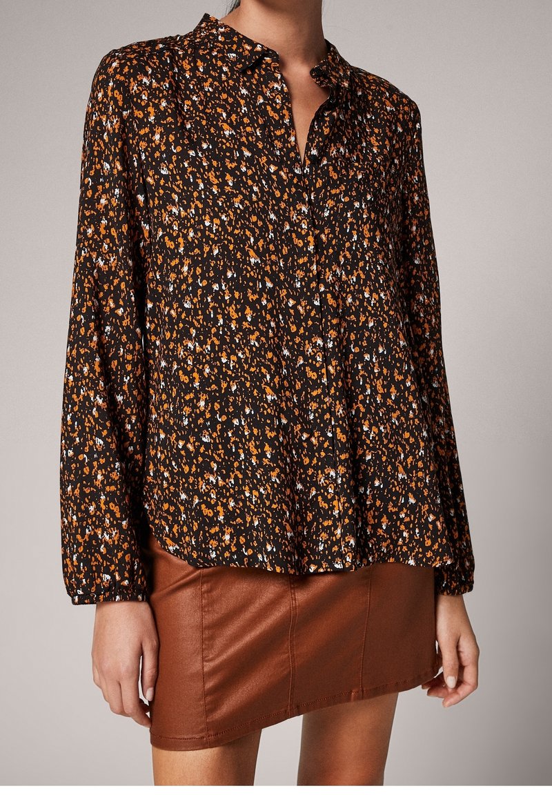 Blouse noire à boutons avec un motif floral orange et bleu. Manches longues avec poignets à boutons, coupe légèrement ample et texture de tissu douce.