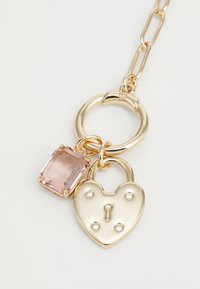 Χρυσού χρώματος με γοητεία που διαθέτει ένα ροζ ορθογώνιο πολύτιμο λίθο και ένα λουκέτο σε σχήμα καρδιάς με προεξέχοντα στοιχεία, και τα δύο κρεμασμένα από μια αλυσίδα �σύνδεσης.